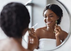 Beautiful african american young woman covered in towel standing in front of mirror and using facial roller and serum, making facial massage after morning shower at home, closeup photo